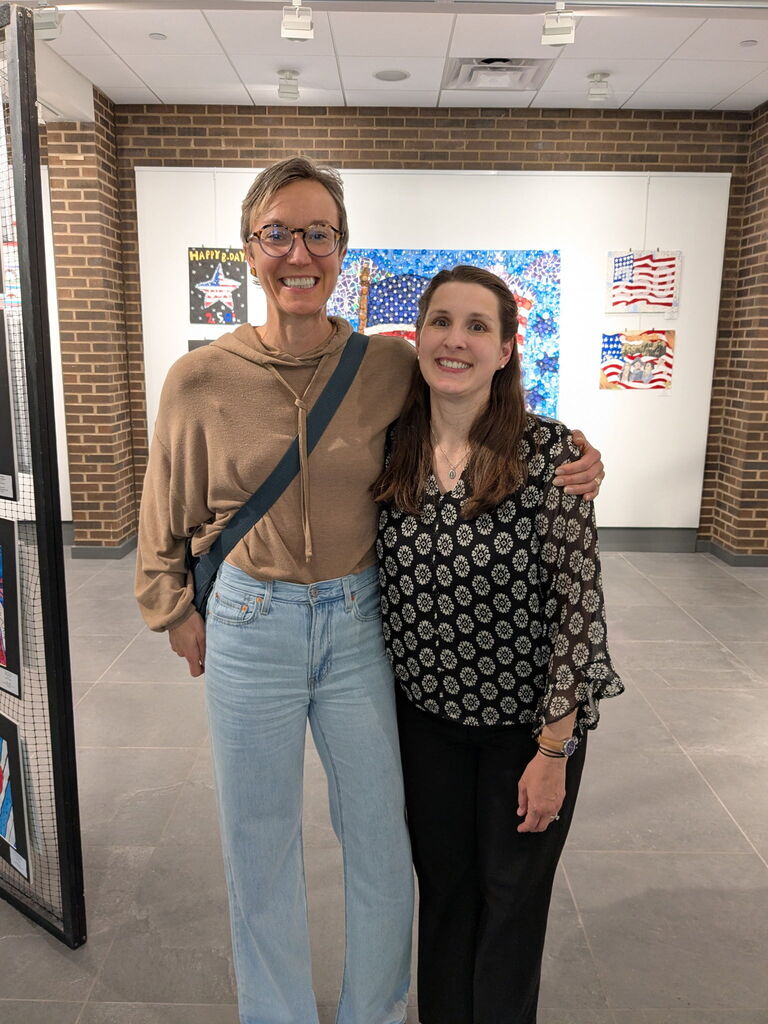 Two adults smile with their arms around each other while standing in front of a student art exhibit featuring patriotic-themed artwork and a large eagle display in the background.