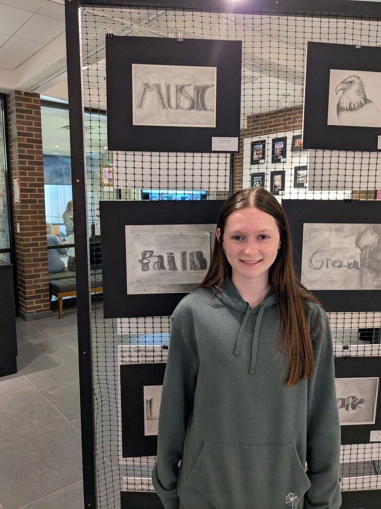 A student poses in front of a display of black-and-white and shaded word art pieces mounted on a wire gallery panel during the student art showcase.