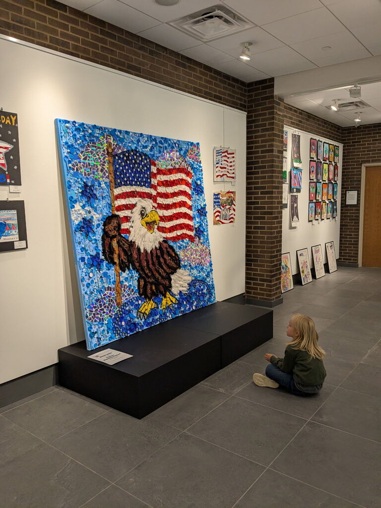 A young student sits on the floor admiring a large eagle and American flag art installation displayed in the center of a student art gallery. Colorful student artwork lines the surrounding walls.