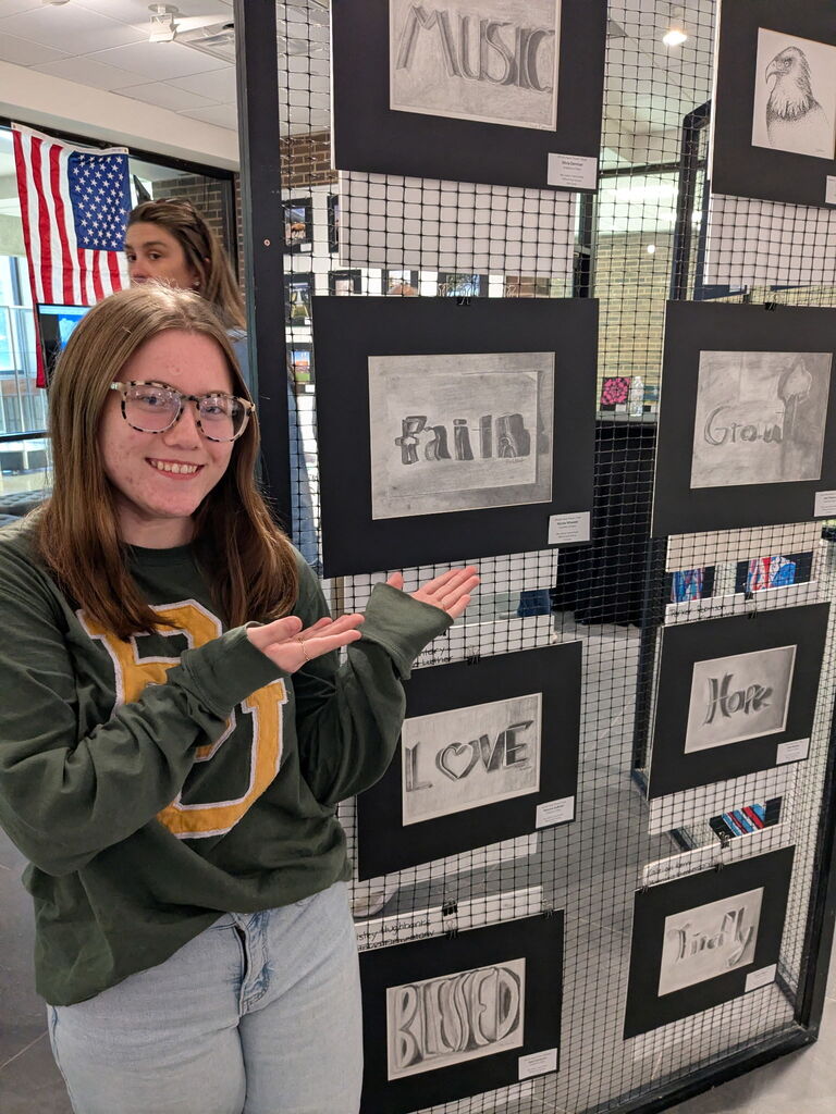 A student wearing glasses gestures toward her displayed artwork mounted on a wire gallery panel featuring stylized shaded words and typography designs.