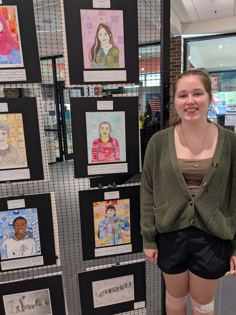 A student smiles beside a gallery display of colorful portrait artwork created by students and mounted on black backing panels.