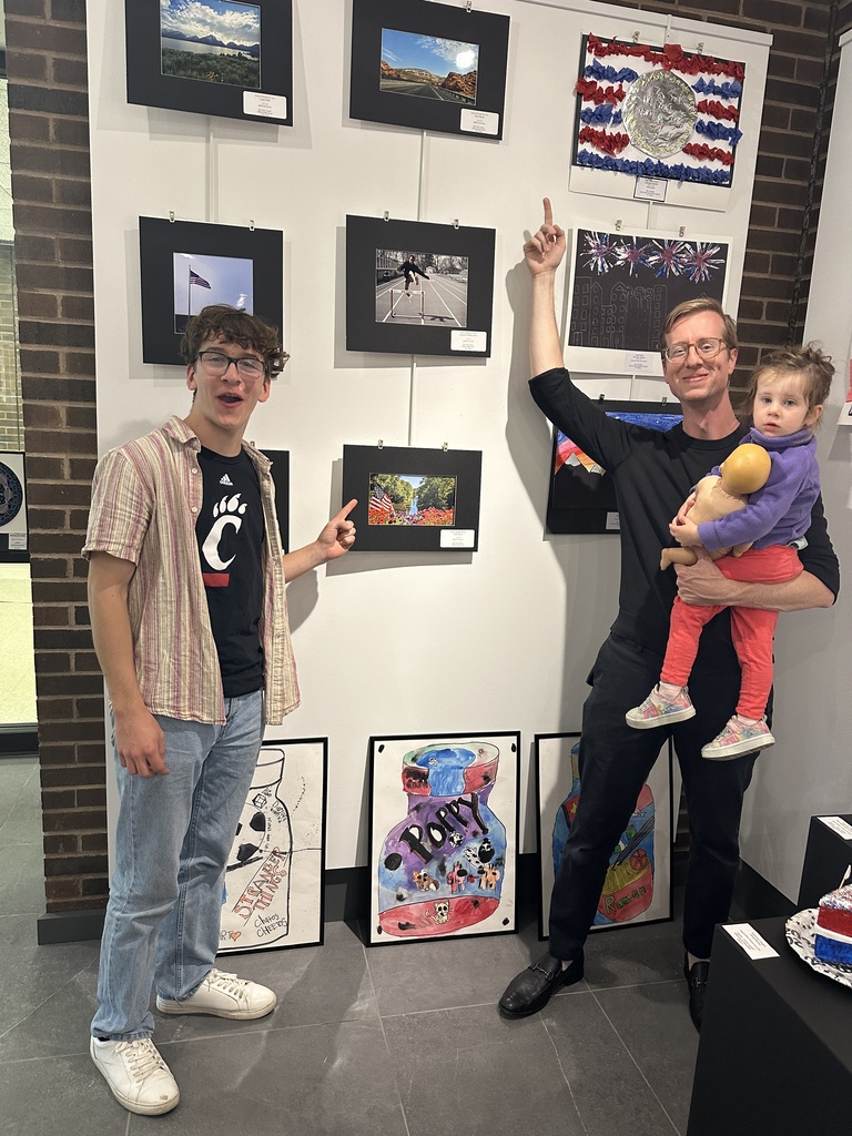 A student and adult point toward student artwork displayed on a gallery wall featuring photography, patriotic-themed pieces, and colorful painted jars while a young child is held nearby.