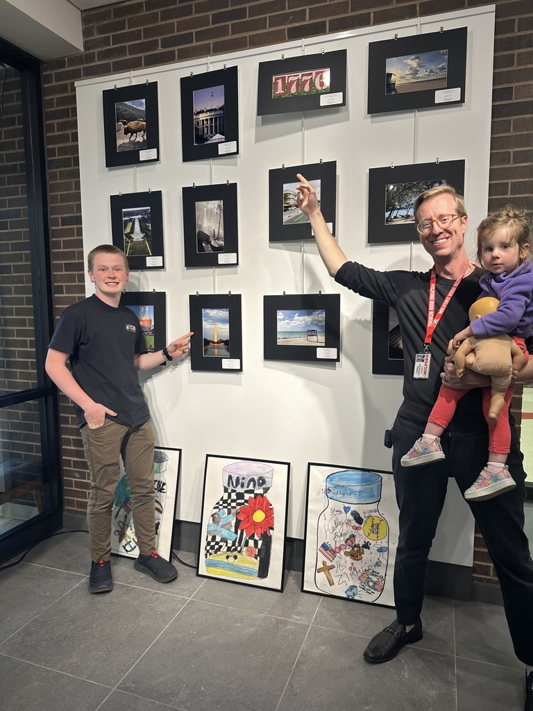 A student and adult pose together while pointing at a photography display wall featuring scenic and patriotic-themed student photographs. A young child is being held beside them.