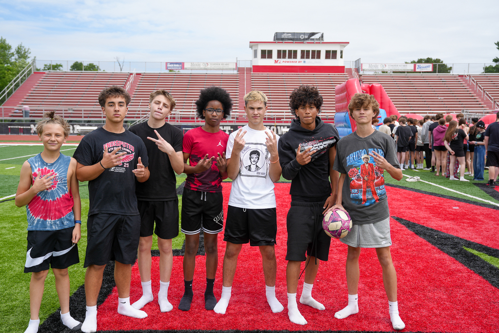 A group of middle school students poses together on the football field during field day with stadium bleachers and inflatables behind them.