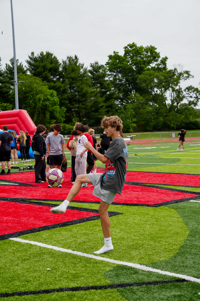 A middle school student balances a soccer ball during a field day activity on the football field while other students gather near inflatable games in the background.