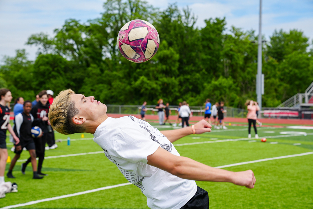 A middle school student heads a soccer ball during a field day activity on the turf field while classmates watch nearby.