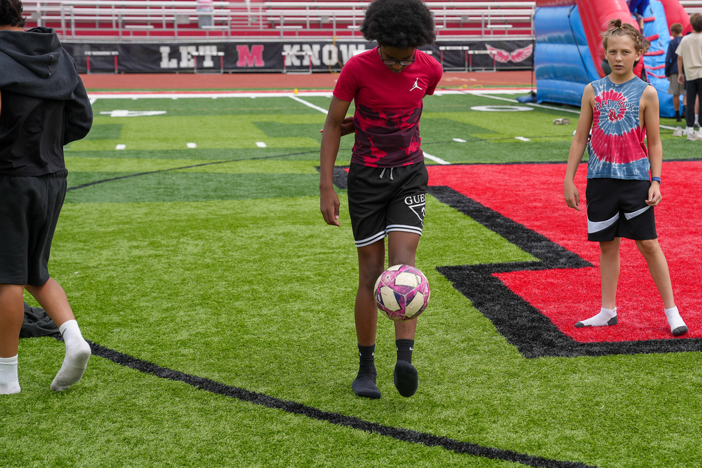 A student juggles a soccer ball on the turf field while classmates stand nearby during middle school field day activities at the stadium.