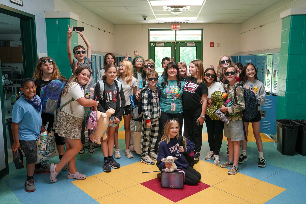 A group of fifth-grade students wearing sunglasses pose for a photo with their teacher inside a school hallway on the last day of school. Many students are smiling, holding backpacks, and making peace signs.