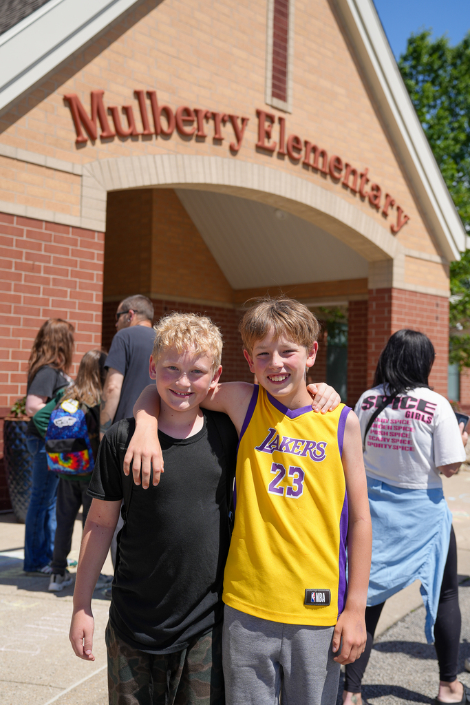 Two students stand outside Mulberry Elementary smiling with their arms around each other on the final day of school. One student wears a yellow Lakers basketball jersey while families and students gather in the background.