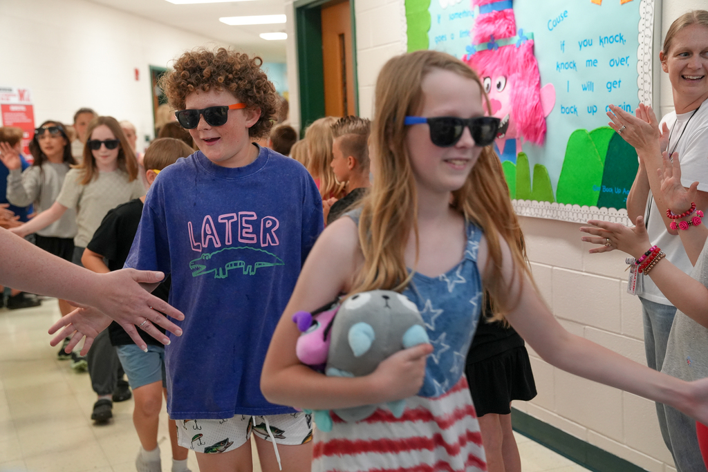 Students wearing sunglasses walk through a school hallway during a fifth-grade walkout while classmates and staff line the hallways clapping and giving high fives. One student carries a stuffed animal while smiling during the celebration.