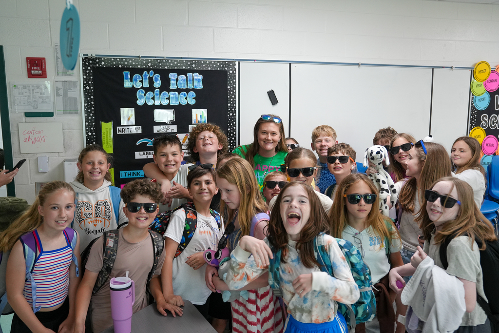 Students wearing sunglasses gather around their teacher inside a classroom on the last day of school while posing for a fun group photo together.