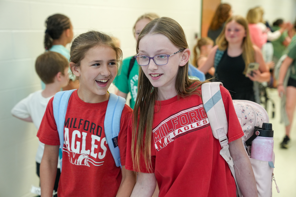 Two students wearing matching red Milford Eagles shirts walk side by side through a school hallway smiling and talking during the final day of school.