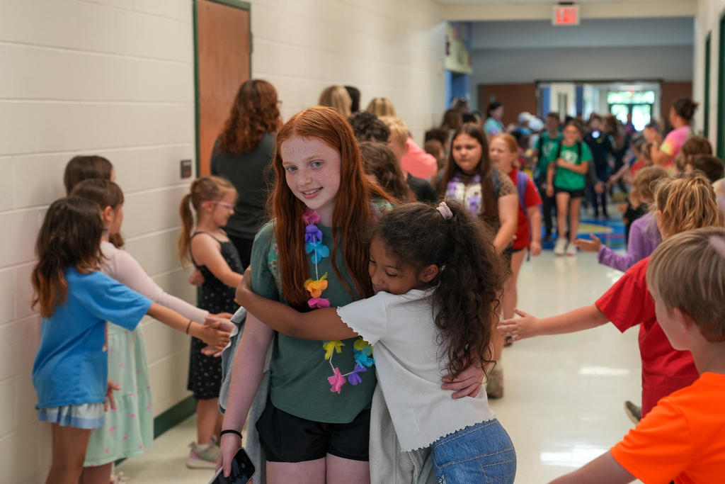 A younger student hugs a fifth-grade student during a hallway walkout celebration while classmates line the hallway giving hugs and high fives.