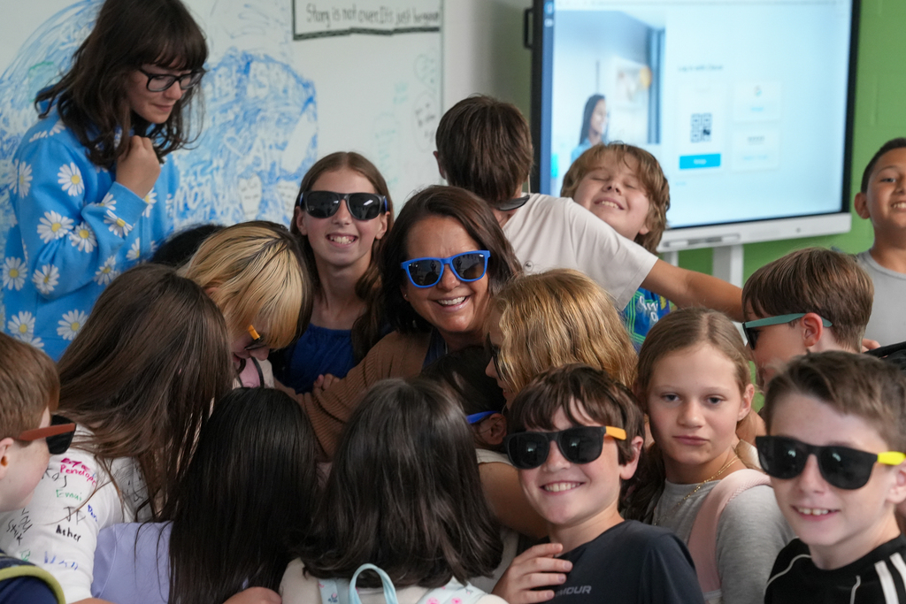 A teacher wearing sunglasses smiles while surrounded by students inside a classroom during an end-of-year celebration. Several students hug together and pose for the camera.