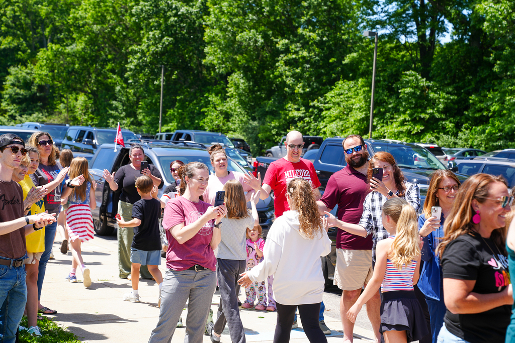 Families, students, and staff gather outside on a sunny day to cheer and celebrate students during an end-of-year walkout. Several parents hold up phones to take photos while students walk past giving high fives.