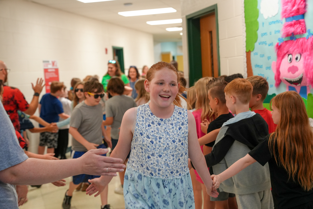 A smiling student in a blue dress walks through a hallway during a fifth-grade walkout as younger students and staff members stand on both sides cheering and high fiving students.