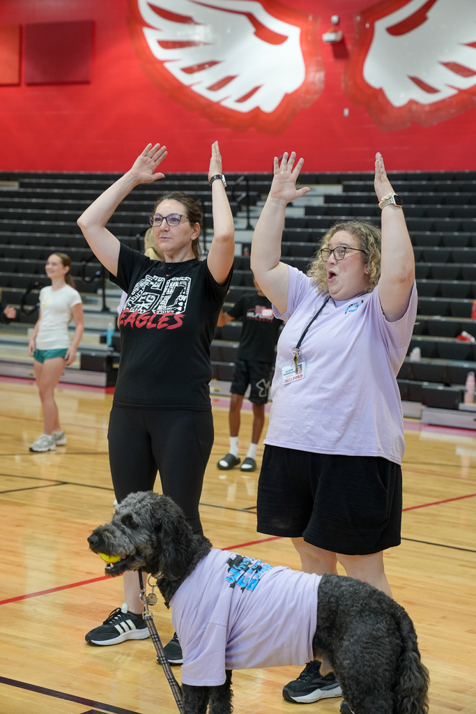 Two staff members raise their hands during a WEB training activity while a therapy dog wearing a purple WEB shirt stands in front of them.