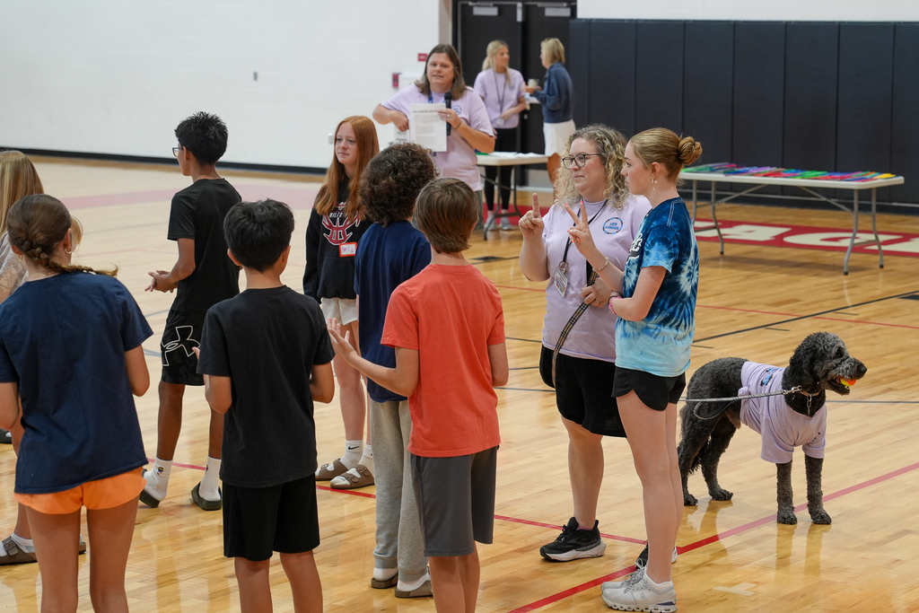 Students and staff members participate in a small group WEB training activity in the gymnasium while a black therapy dog wearing a purple WEB shirt stands nearby.