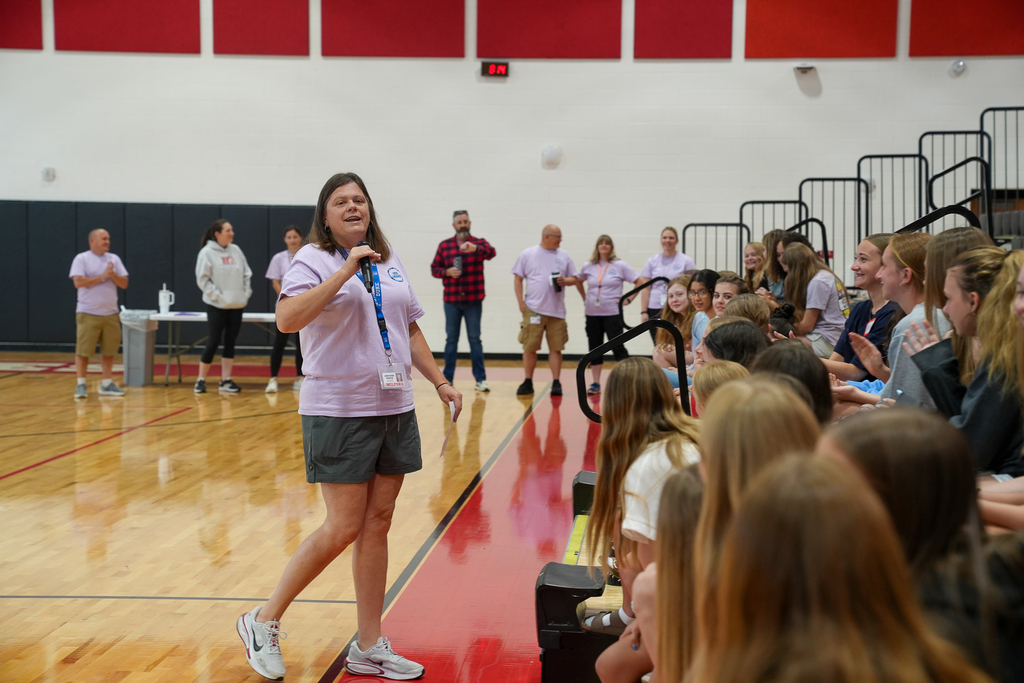 Students participate in WEB (Where Everybody Belongs) training inside the Milford Middle School gymnasium while a staff member speaks into a microphone to seated students in the bleachers. Additional staff members stand in the background observing the activity.