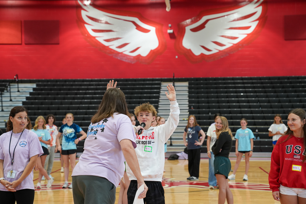 A student wearing a Milford Lacrosse sweatshirt raises his hand while participating in a WEB leadership activity in front of classmates and staff members.