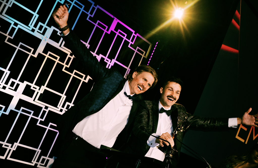 A photo shows two men in tuxedos celebrating onstage while holding an Emmy award trophy. One man raises his arm in excitement as both smile toward the audience. Bright stage lighting and colorful geometric backdrop designs create a dramatic awards-show atmosphere behind them.
