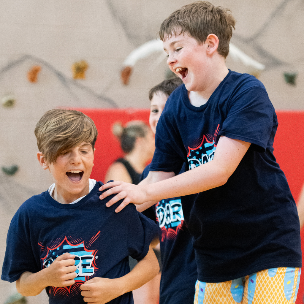 In a gymnasium, two students wearing D.A.R.E. shirts laugh and celebrate during an indoor game while classmates watch nearby.