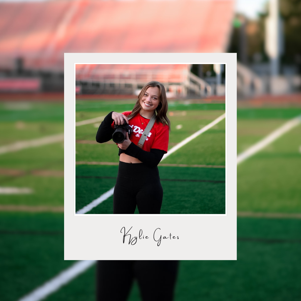 A smiling young woman stands on a football field holding a professional camera. She is wearing a red shirt over a black long-sleeve top and black pants, with a camera strap around her neck. The image is styled like a Polaroid photograph, with her portrait framed in a white border and the name “Kylie Gates” written in script below the photo. The background features blurred stadium bleachers and the football field, creating a shallow depth-of-field effect that keeps the focus on Kylie and her camera.