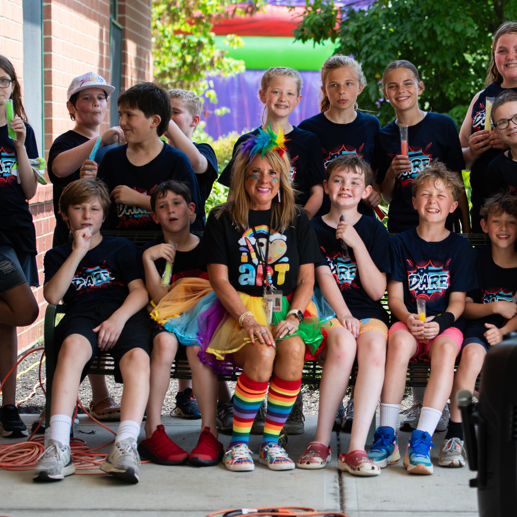 A group of students wearing D.A.R.E. shirts gather around a staff member dressed in a colorful rainbow-themed outfit, posing together for a group photo.