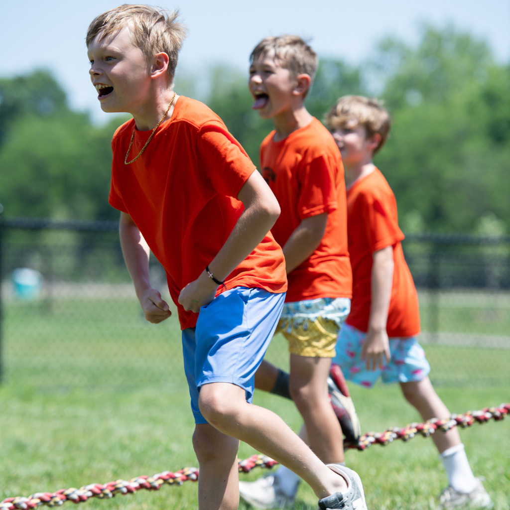 Three students in orange shirts celebrate a tug of war victory , cheering and laughing in a grassy field