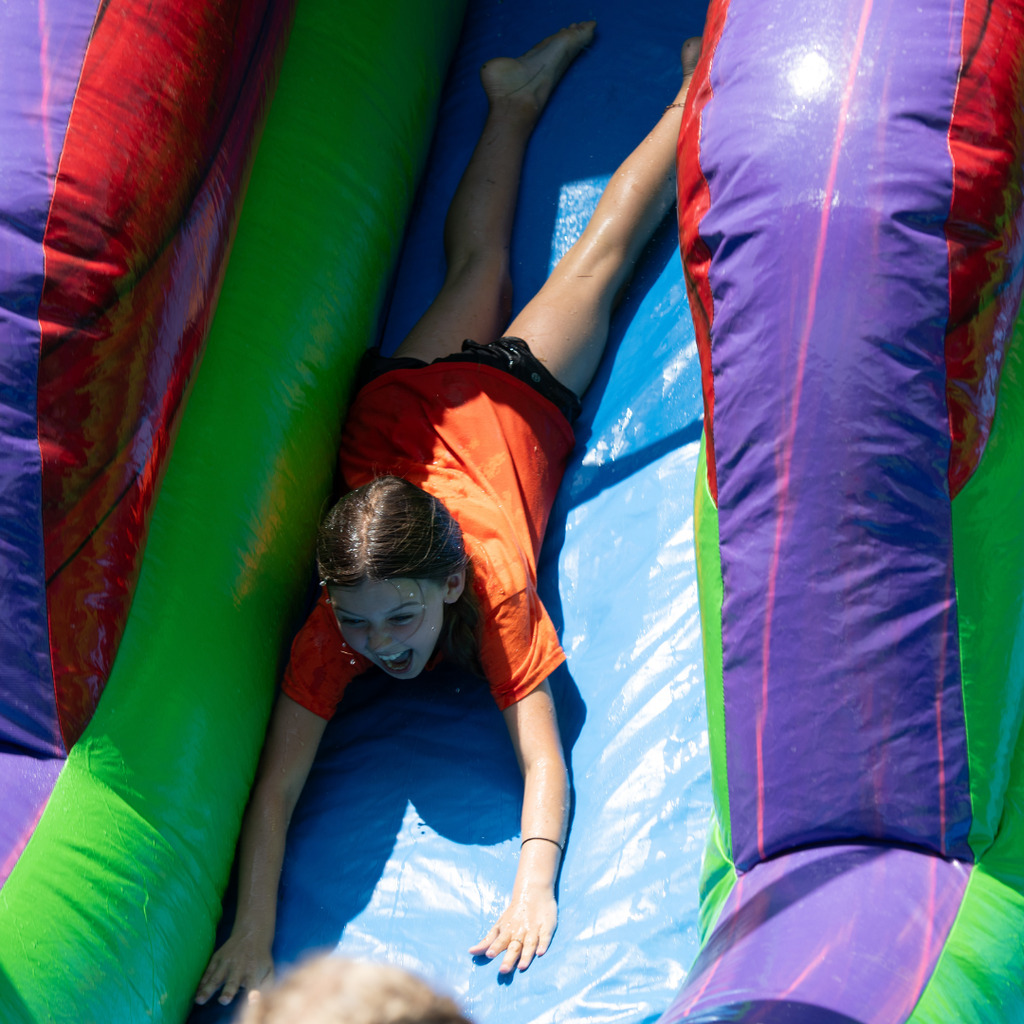 In one image, a student in an orange shirt laughs while sliding headfirst down a colorful inflatable water slide.