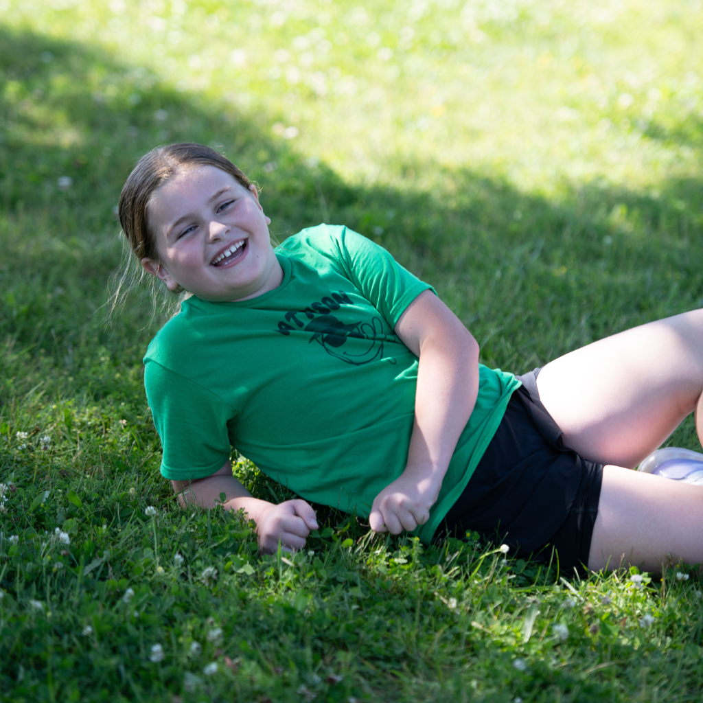A student in a green Pattison shirt relaxes in the grass, smiling after participating in activities.