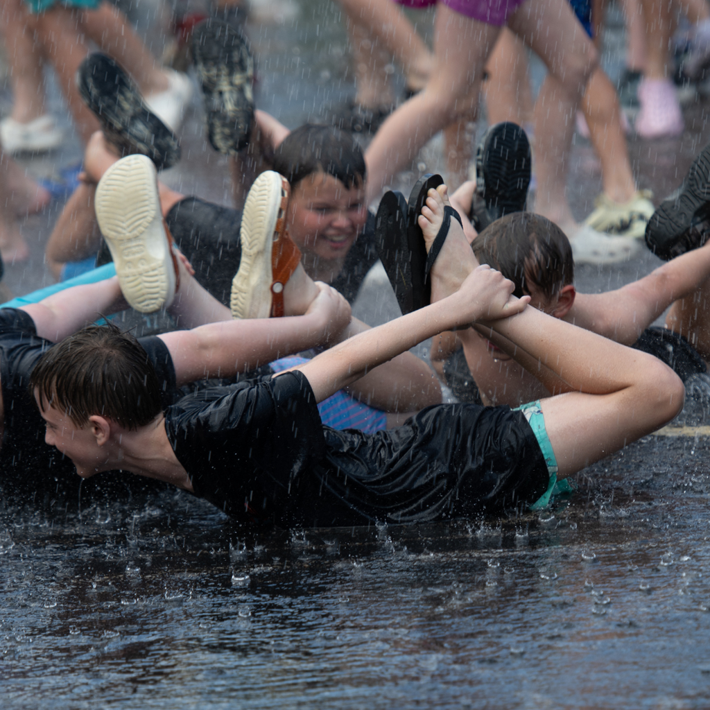 Several students laugh and splash through a wet outdoor activity during a school field day event. In the foreground, two students lie on their stomachs on a slick, water-covered surface while gripping each other's arms as they slide through puddles. Water sprays through the air around them, and their clothing is soaked from the activity. Additional students can be seen in the background participating in the game, with legs and feet visible as they run and splash through the water. The image captures the excitement, energy, and teamwork of students enjoying a summer-themed field day challenge.