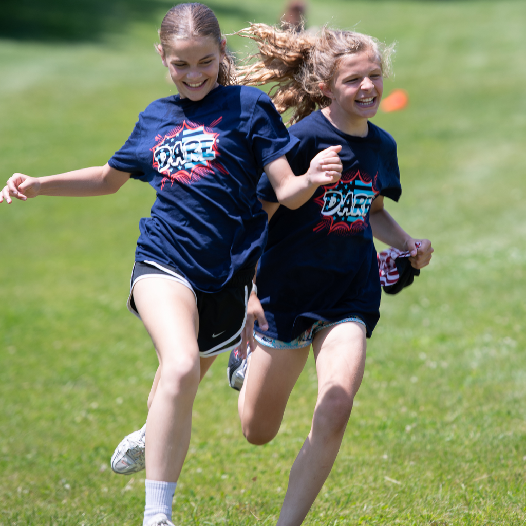 Two students in navy D.A.R.E. shirts sprint side by side across the grass, smiling as they compete in a race.