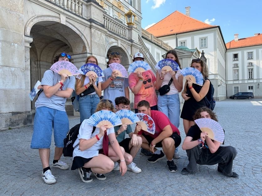 A group of students poses in a courtyard of a historic European building, holding decorative fans while smiling for a group photo.