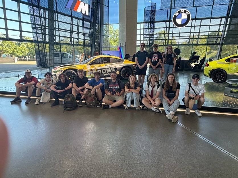A group of students poses together inside a BMW museum, sitting and standing in front of displayed race cars and automotive exhibits.