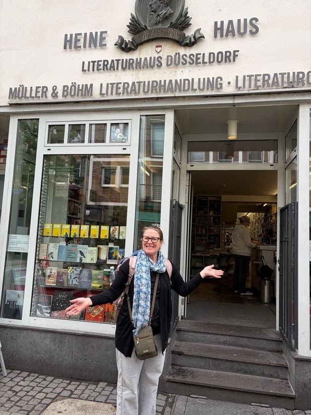 A woman stands outside the entrance of Heine Haus Literaturhaus Düsseldorf, smiling and gesturing toward the bookstore entrance in a welcoming pose.