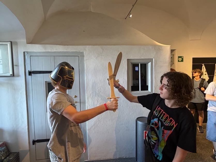 Two students participate in a hands-on historical activity, playfully fencing with wooden swords while wearing protective helmets inside a museum-style exhibit space.
