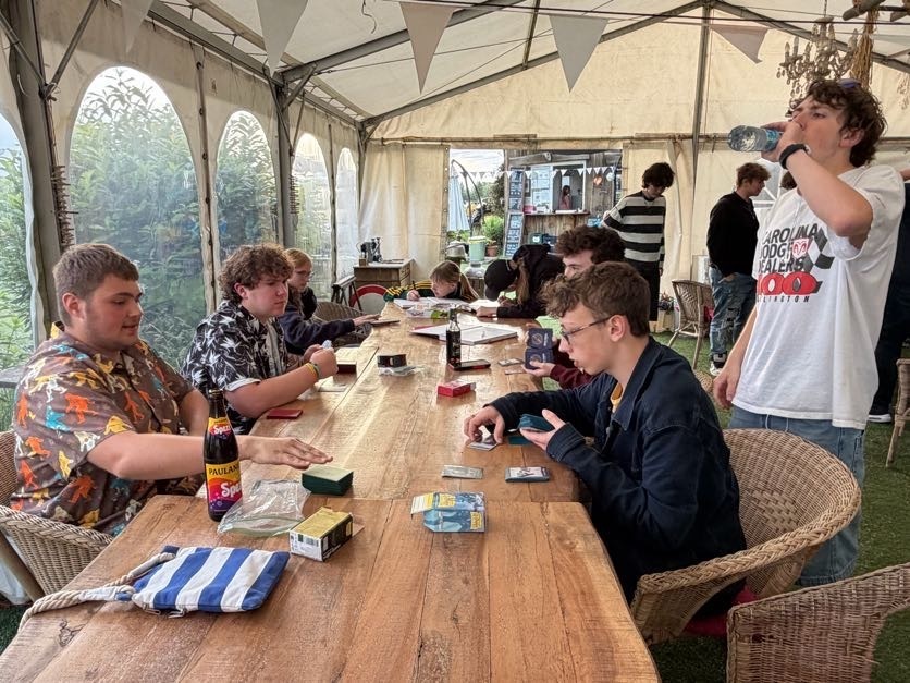 A group of students sits around a long wooden table inside a tented outdoor space, playing cards and socializing in a relaxed setting.