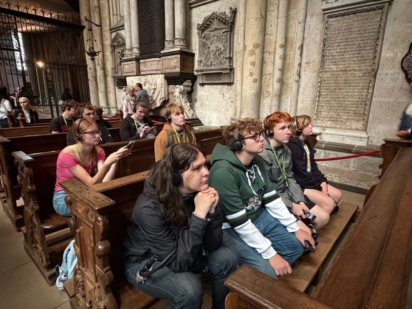 A group of students sits quietly in wooden pews inside a large historic church or cathedral, wearing headphones as they listen to an audio-guided tour.