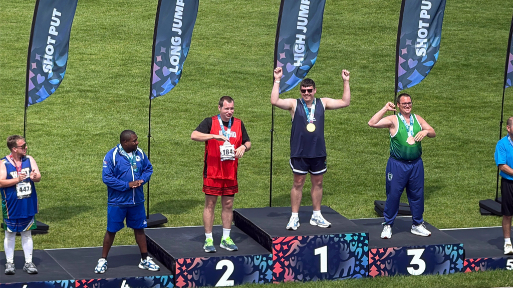 Jackson Witsken stands atop the first-place platform of an outdoor awards podium, wearing a gold medal and raising both arms in celebration. Other athletes stand on the surrounding podium platforms.