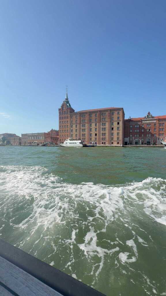 View from a boat across the Venetian Lagoon, showing historic brick waterfront buildings under a clear blue sky with waves in the foreground.