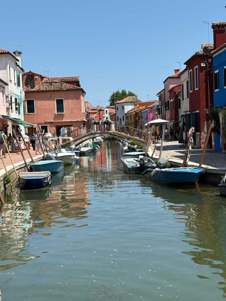 Colorful buildings line a narrow canal in Murano, Italy, with small boats docked along the water and a pedestrian bridge in the distance.