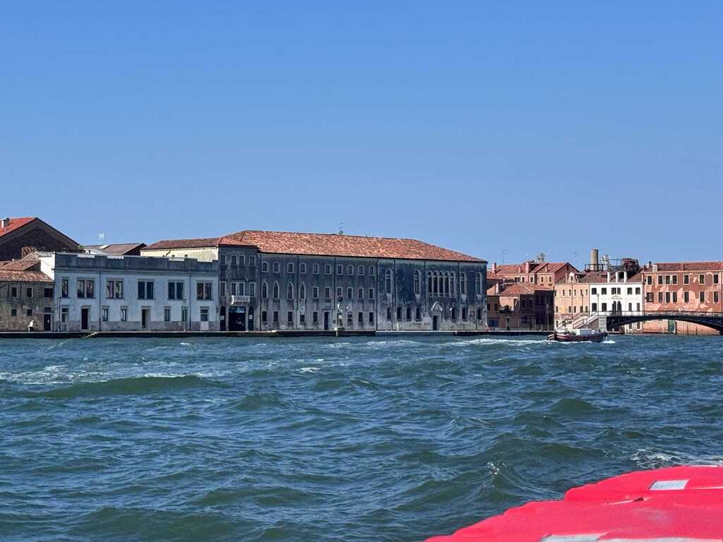 Historic waterfront buildings and a small bridge along the Venetian Lagoon, with choppy blue-green water in the foreground.