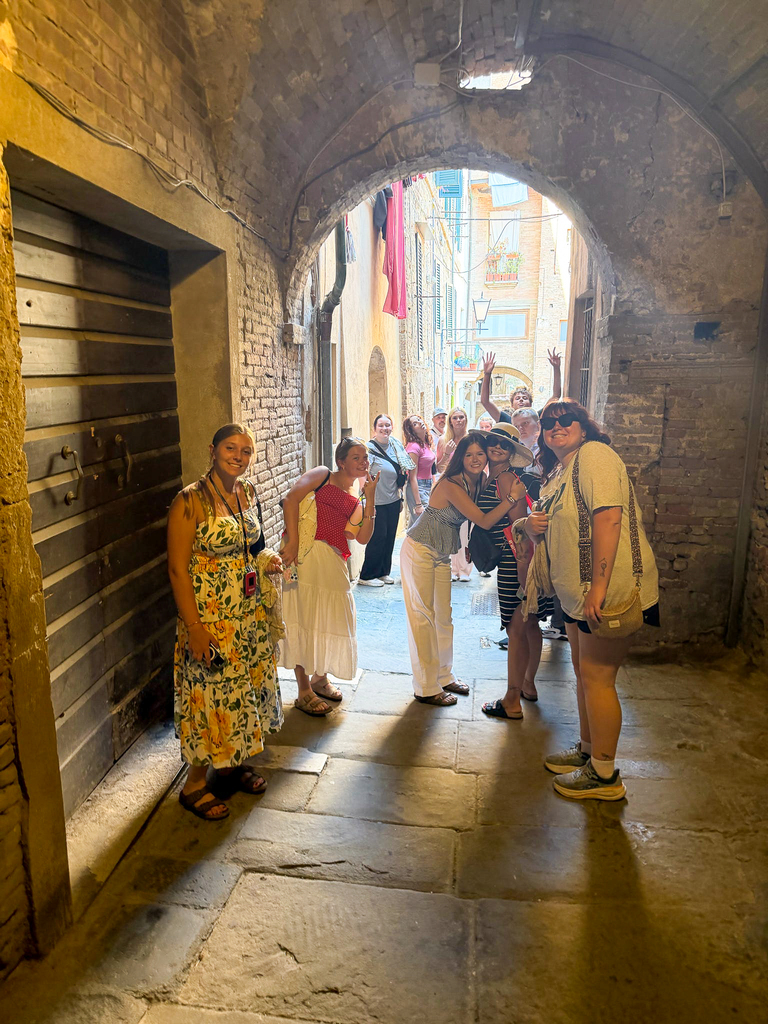 A group of Milford High School students and adult chaperones pose together beneath a large stone archway in a narrow historic street in Siena, Italy.