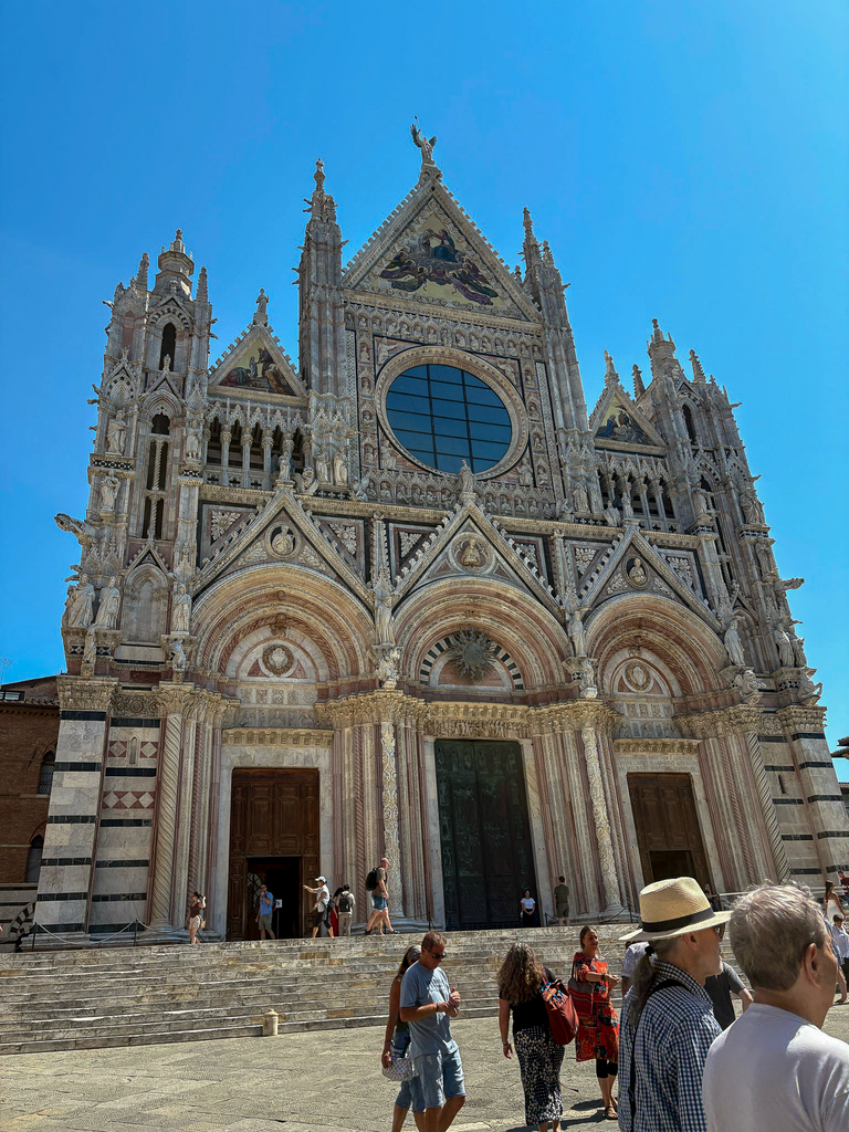 The ornate Gothic façade of the Siena Cathedral rises against a bright blue sky, with visitors gathered on the steps below.