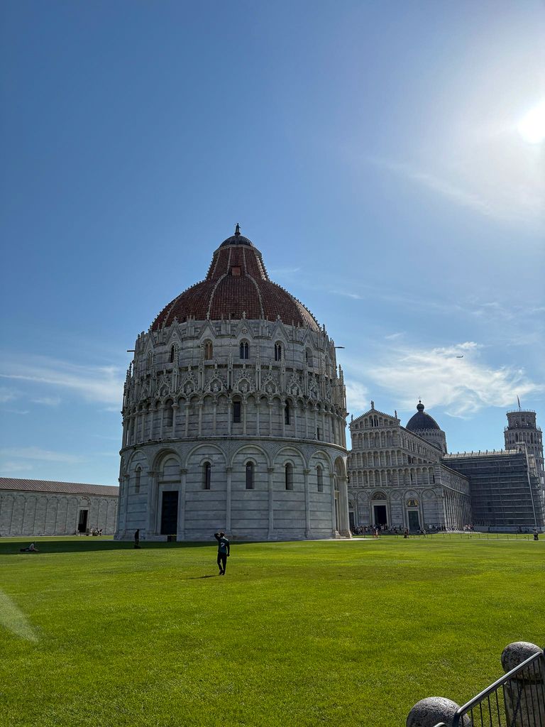 The Pisa Baptistery stands on a large green lawn in the Piazza dei Miracoli, with the Pisa Cathedral and Leaning Tower visible in the background.