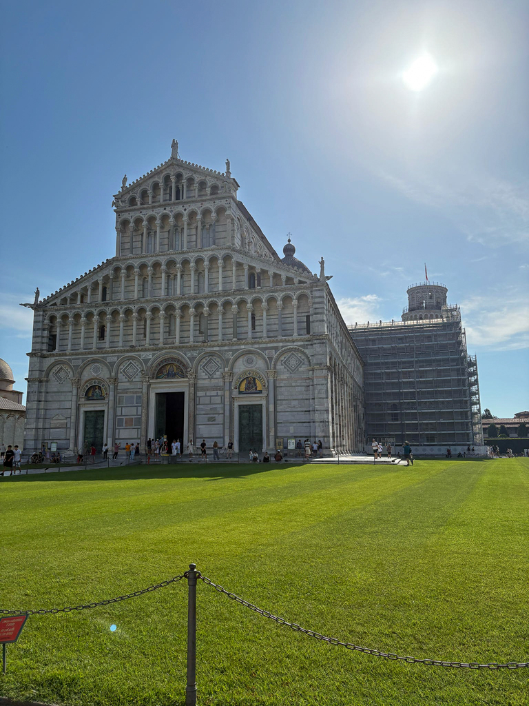 The Pisa Cathedral stands beneath a bright blue sky, with the Leaning Tower of Pisa partially visible behind it.