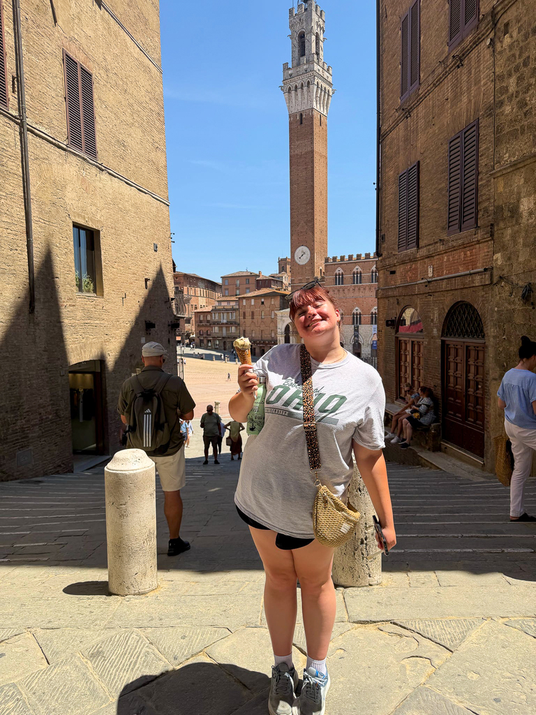 A Milford High School traveler smiles while holding a gelato cone on a stone street overlooking Piazza del Campo, with Siena’s Torre del Mangia rising in the background.