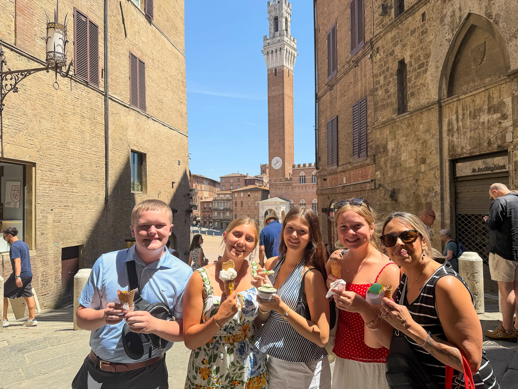 Five Milford travelers pose together with gelato on a historic stone street in Siena, with the Torre del Mangia visible behind them.