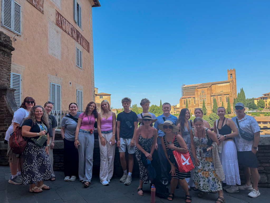 A large group of Milford High School students and adult chaperones pose together at a scenic overlook in Siena, with historic buildings and a large brick church visible in the background.
